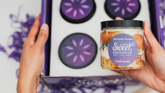Person holding a jar of a Sweet Encounter gluten free cake with a box of similar jars in the background