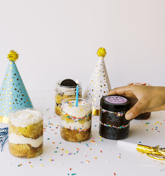 Small cake jars with candles and party hats on a white background