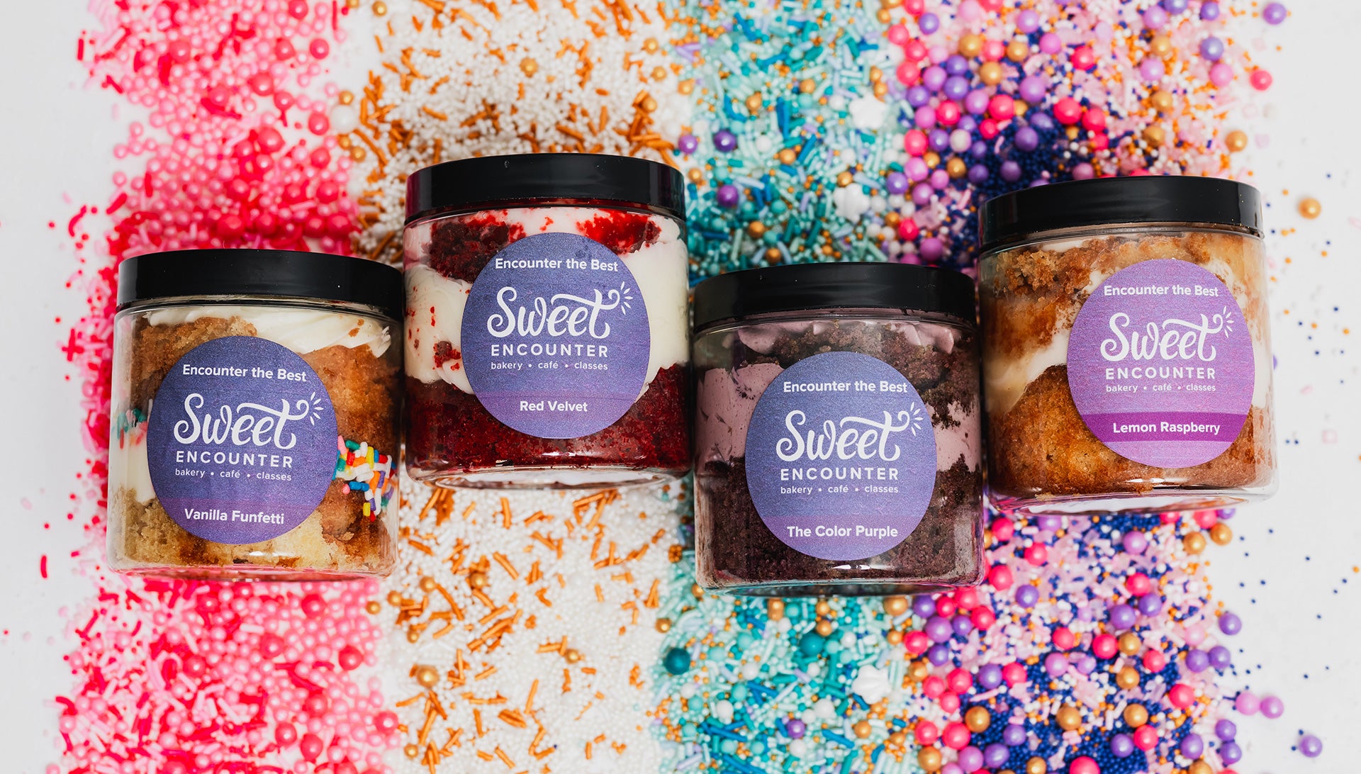 Five jars of gluten free cakes with 'Sweet Encounter' labels on a colorful sprinkles background